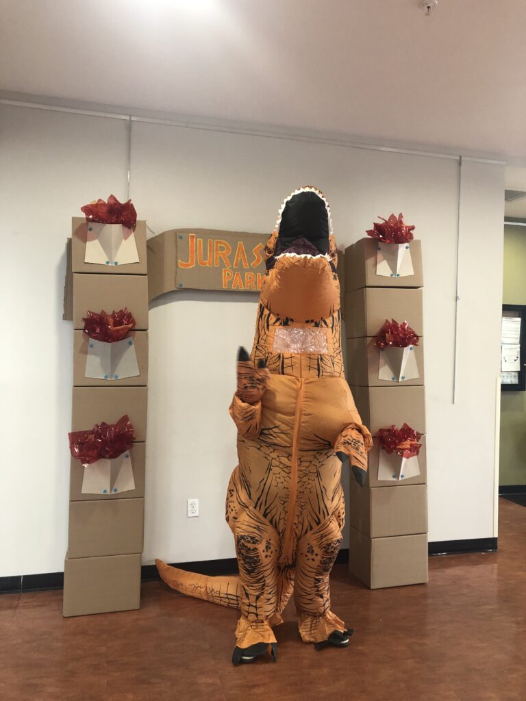 A person in an orange inflatable dinosaur suit waves with one hand in front of a cardboard arch with a painted sign that says “Jurassic Park.”
