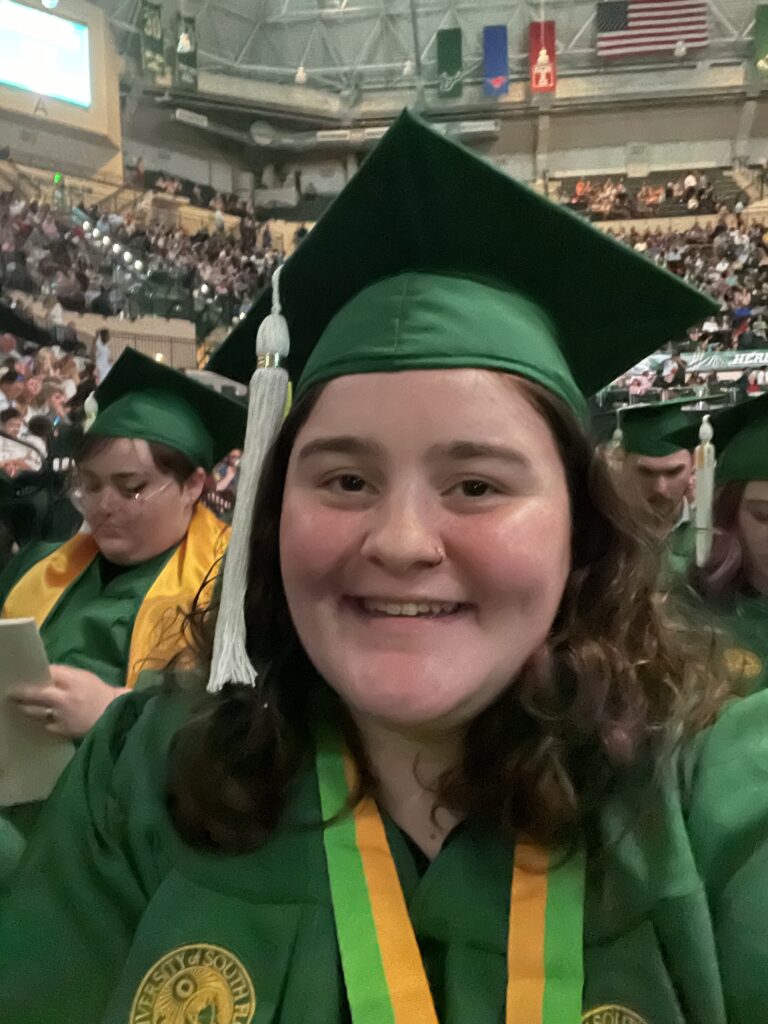 Depicted is a girl with brown, curly hair and brown eyes dressed in graduation commencement garb. The picture is from the chest up, selfie style, and she is smiling. The gown and cap are a solid green. The university's emblem is half-shown from the top in gold on a chest pocket. The tassel on the cap is white. There is a gold and yellow lanyard around her neck. In the background are other graduates and people seated farther back around the stadium seats.