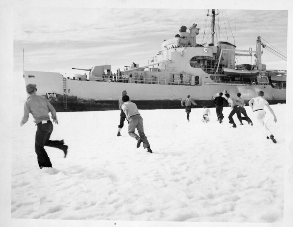 Members of a 1947 expedition to Antarctica play in the snow and ice. A Navy icebreaker ship sits in the background from which onlookers observe the playtime