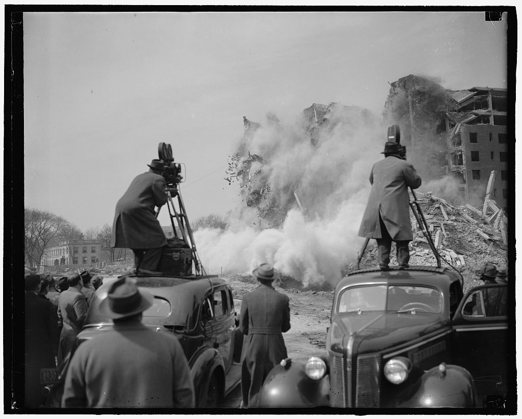 Cameramen stand atop vehicles to shoot footage of a building's demolition in 1940. Black and White