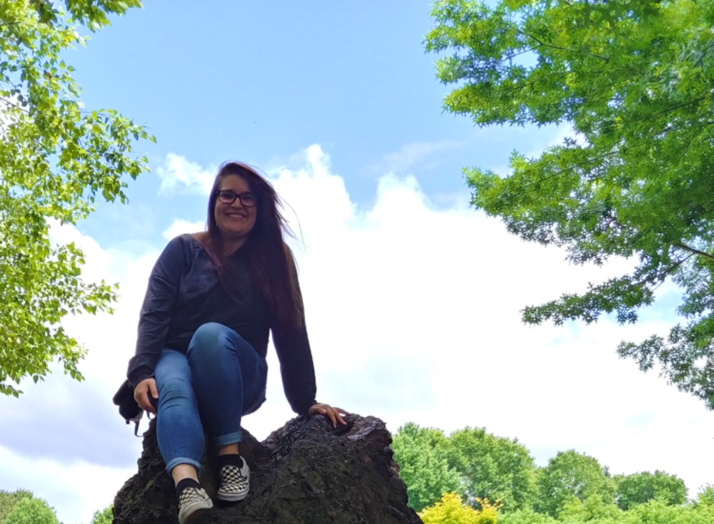 A young woman sits smiling on a large rock, framed by green trees and a bright blue sky.